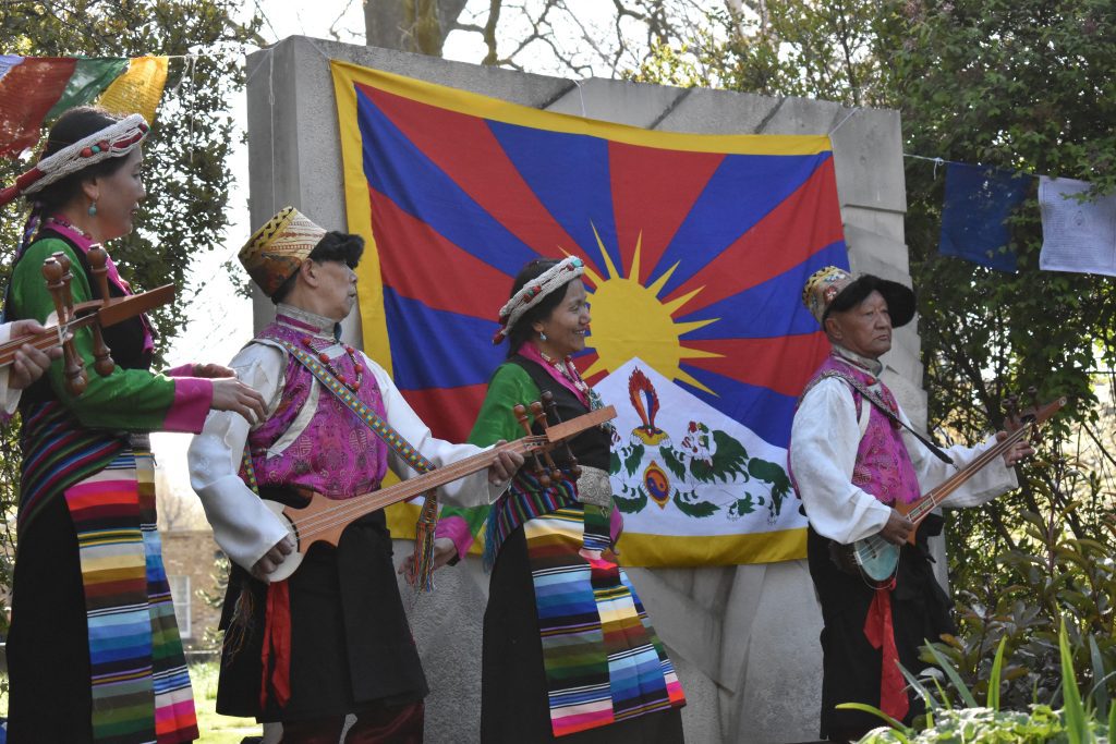 Tibetan community members, dressed in Traditional outfits, dance and play instruments in front of the Tibetan flag.