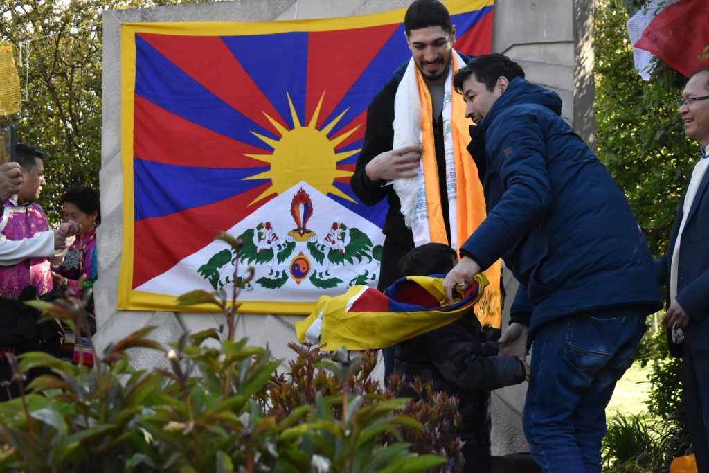 A Tibetan child presents Enes Kanter Freedom with a khata as his father stands by smiling.