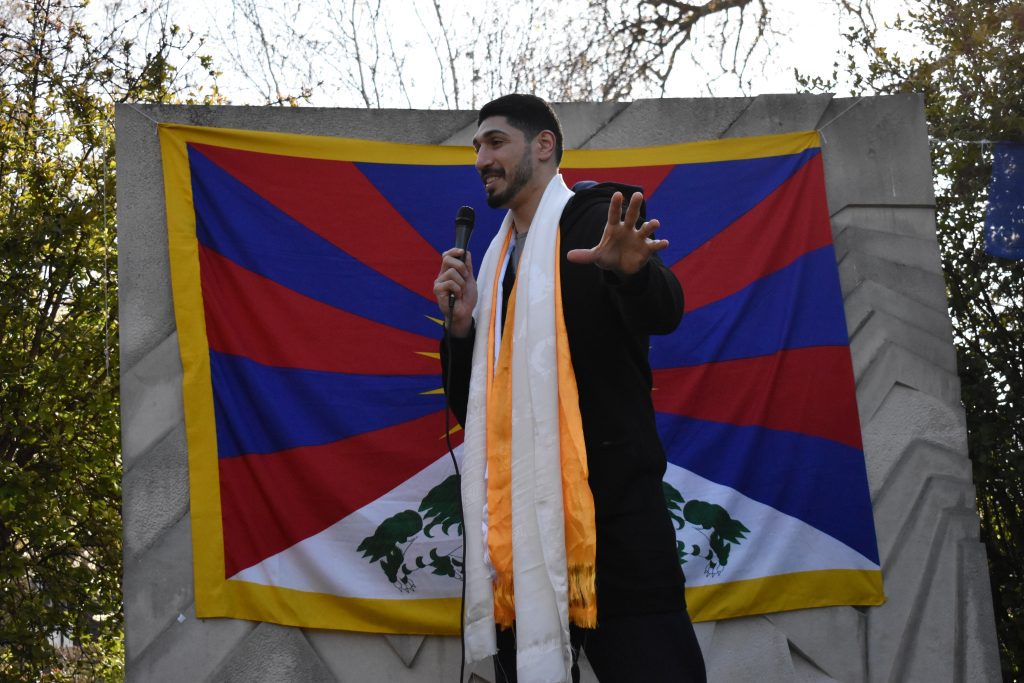 Enes Kanter Freedom stands in front of the Tibetan flag, gesturing with an open hand and smiling as he speaks.